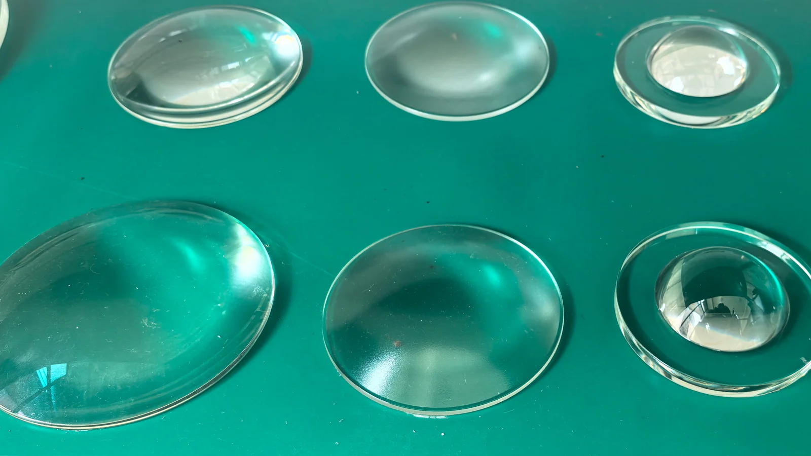 Six circular curved glass components with different edge profiles and visible surface finishes on a green surface