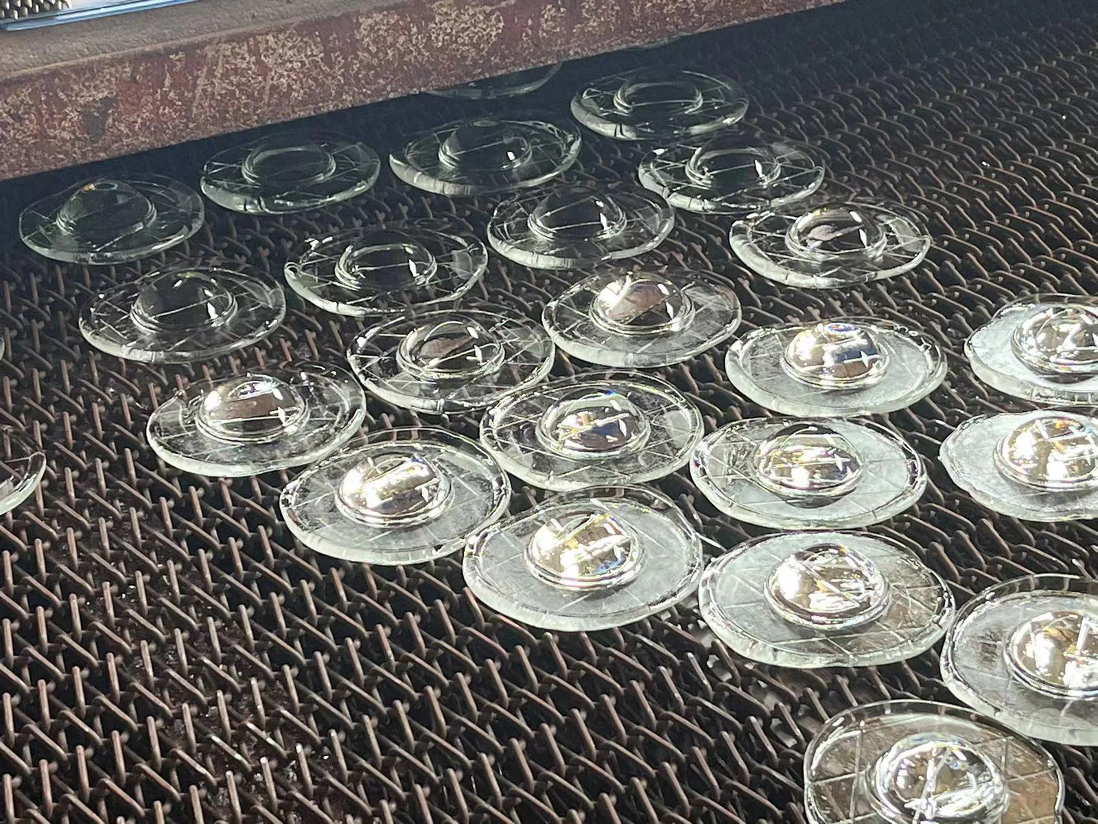 Clear circular glass components arranged on a metal mesh production conveyor