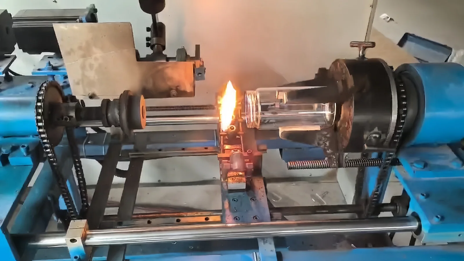 A clear glass component held in a glassworking lathe, with a local flame near its opening and a tool opposite it.