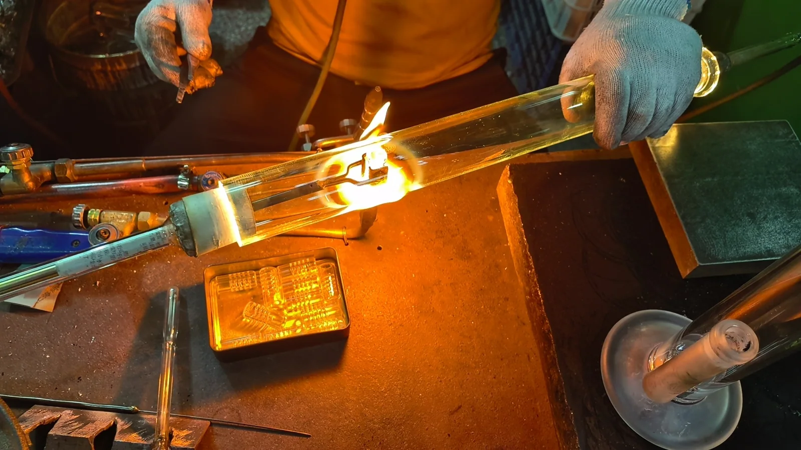 Gloved hands holding a clear glass tube across a local flame, with a hand tool above the working area.